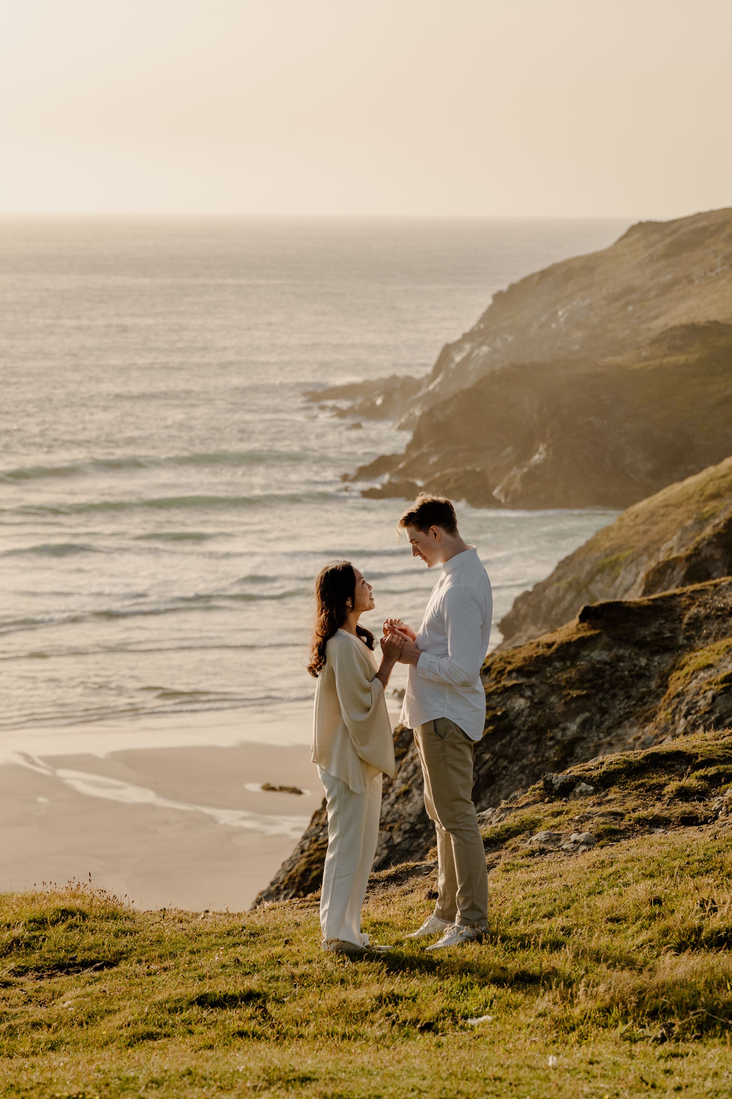 Couple holding hands on coastal cliffs at golden hour with ocean waves and rocky headland behind them.