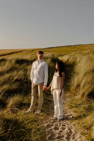 Couple holding hands on sandy path through tall grass dunes in warm evening sunlight.