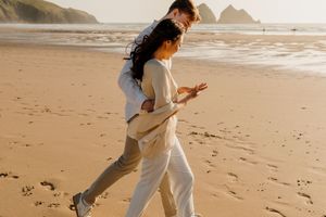 Couple walking hand in hand on a sandy beach at golden hour, dressed in white outfits with ocean waves behind them.