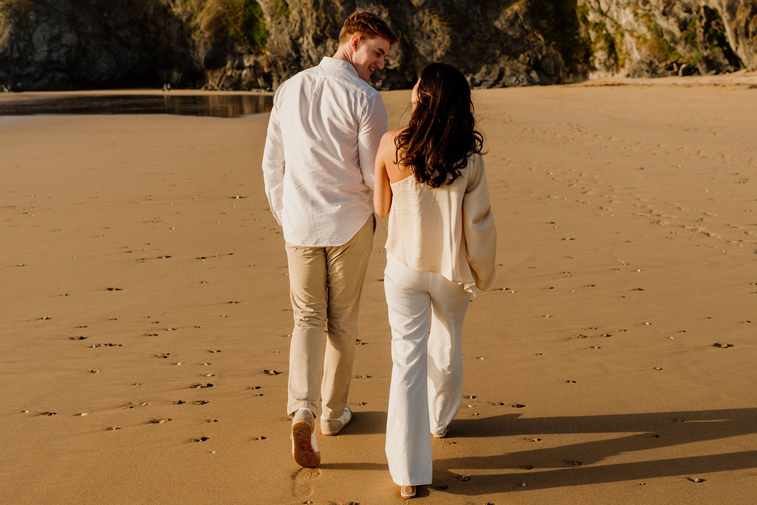 Couple walking hand in hand on a sandy beach at golden hour, dressed in white and beige outfits.