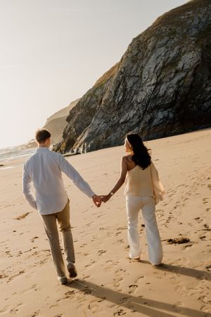 Couple holding hands walking on sandy beach with rocky cliffs in background during golden hour sunset.