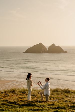 Man proposes to woman on one knee at a scenic beach with rocky ocean islands at golden sunset.