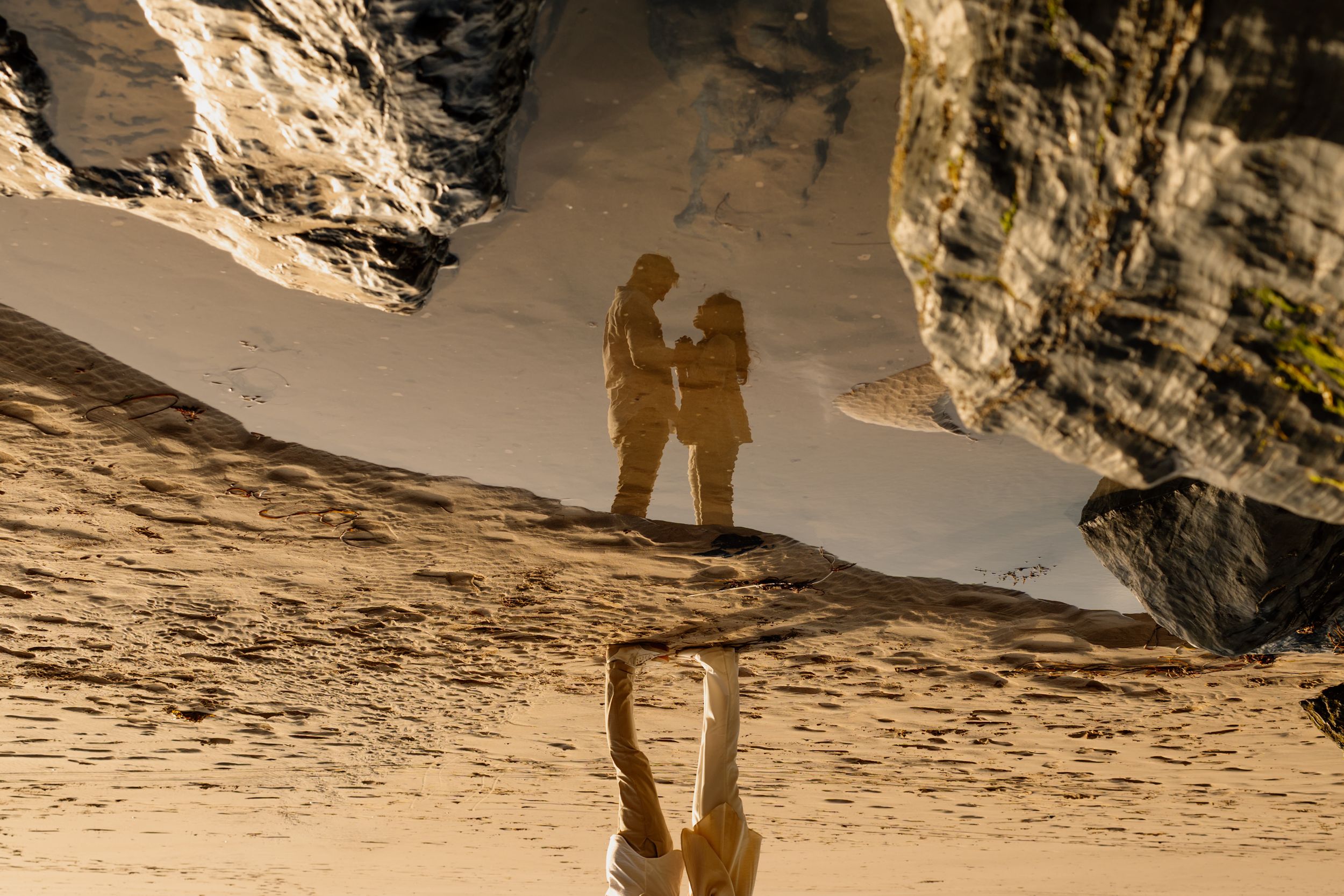 Couple standing on sandy beach with rocky cliffs, reflected in wet sand below, captured in warm golden light.