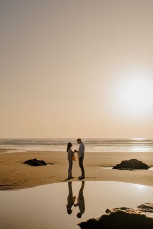 Couple holding hands on a golden sandy beach at sunset, their reflections visible in a tidal pool with ocean waves behind.