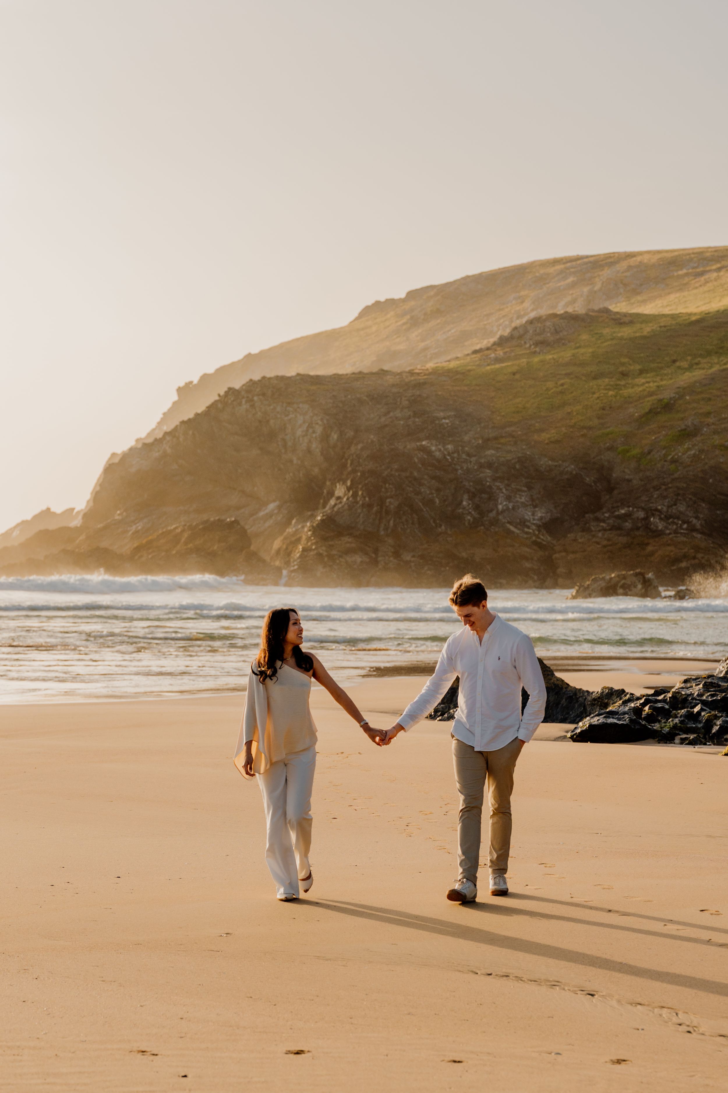 Smiling couple walking hand in hand on sandy beach with dramatic green coastal cliffs at golden hour.