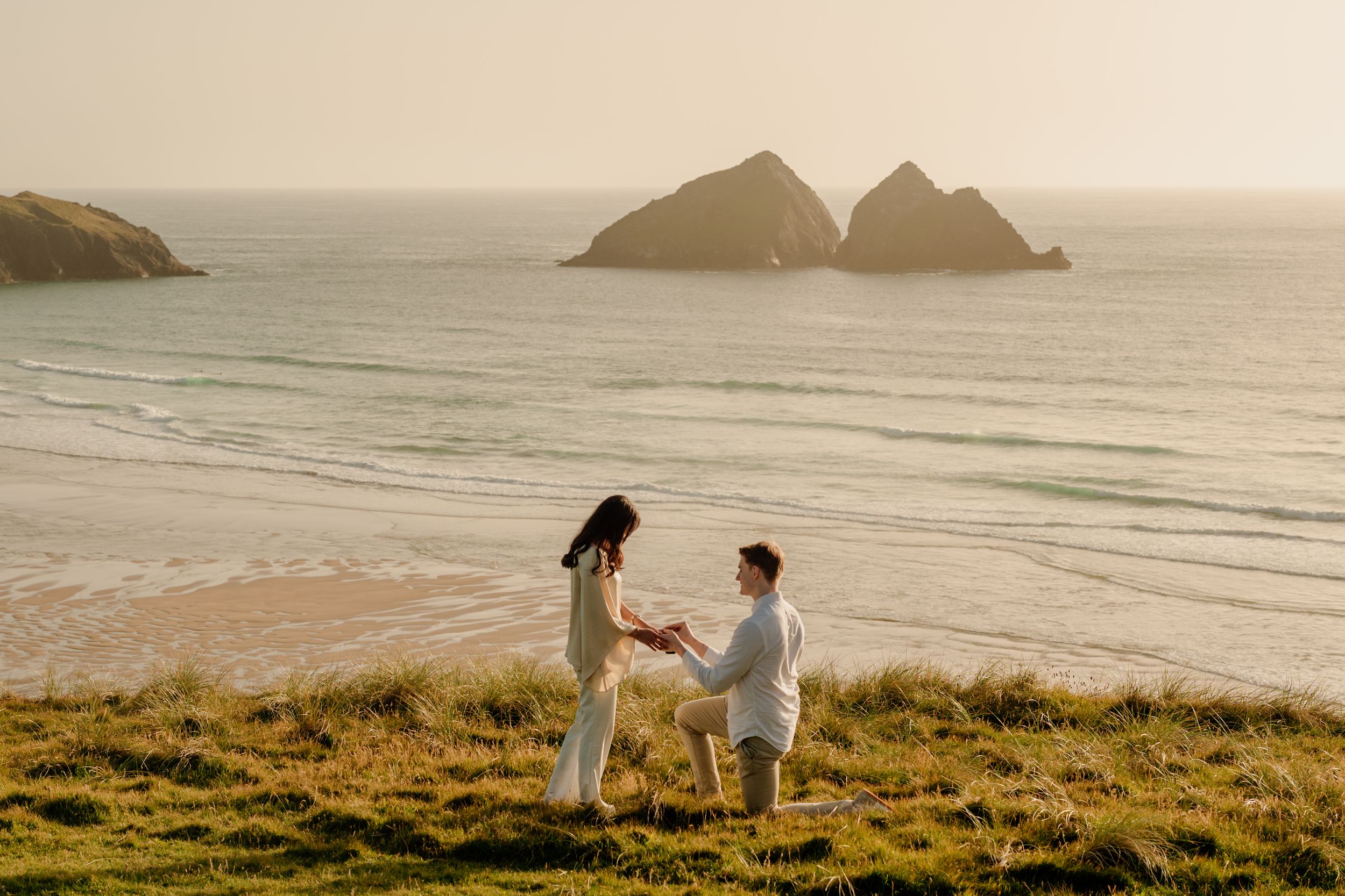 A man proposes on one knee to a woman on a coastal cliff overlooking a sandy beach and rocky islands at sunset.