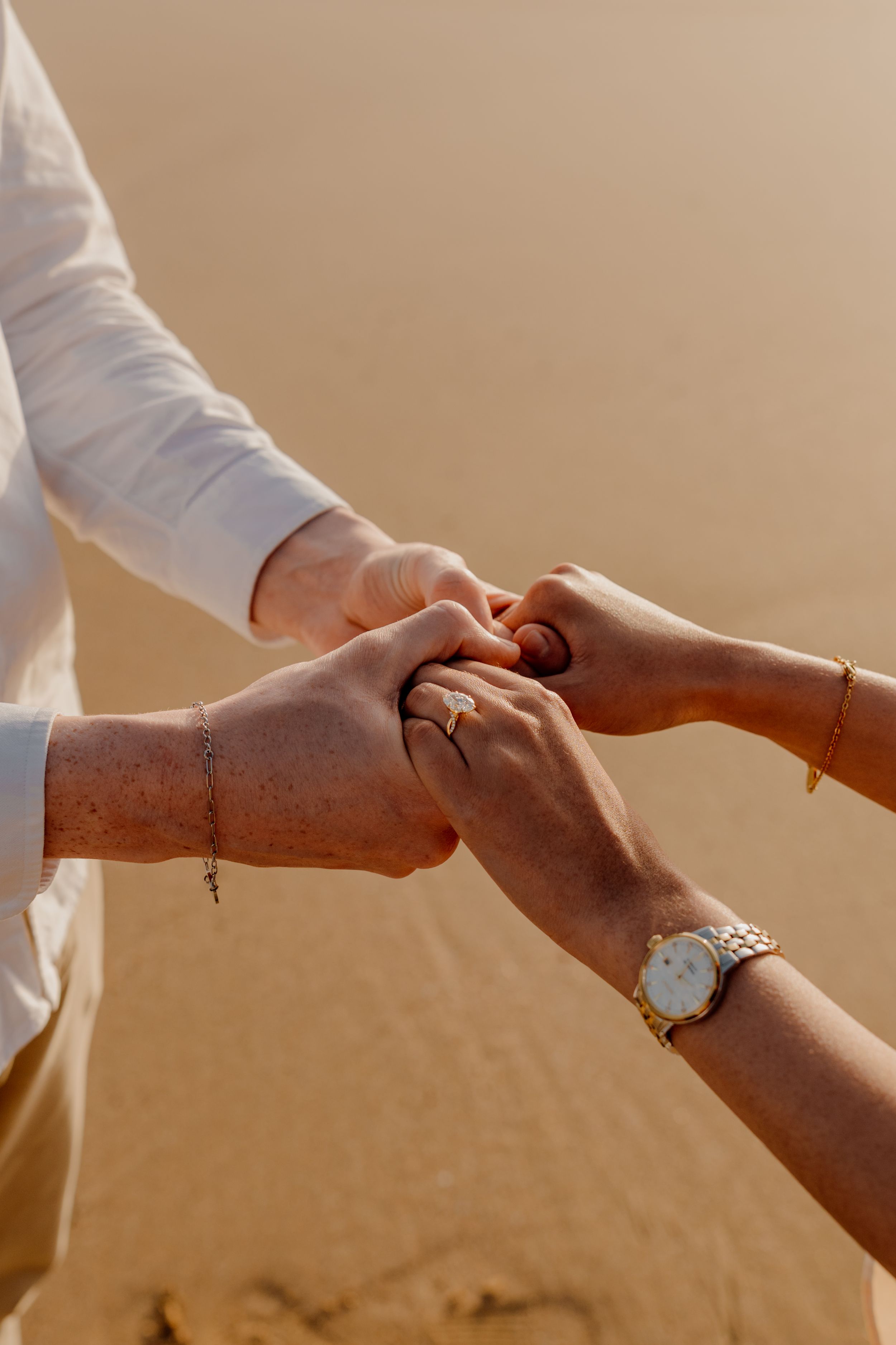 Close-up of a couple holding hands on the beach, woman wearing a ring and gold watch, man in a white shirt with a bracelet.