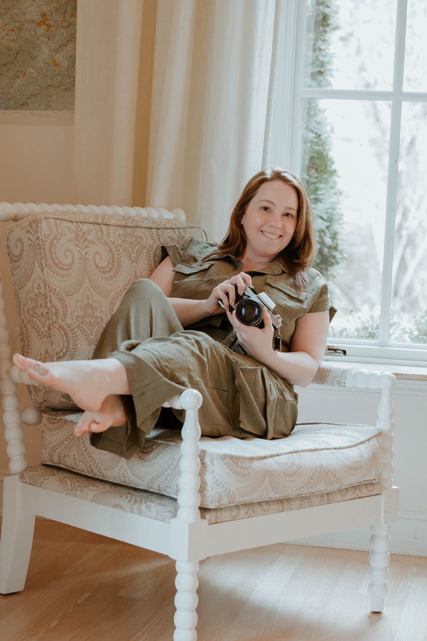 Smiling woman in olive jumpsuit relaxing in white armchair holding a camera near a bright window.