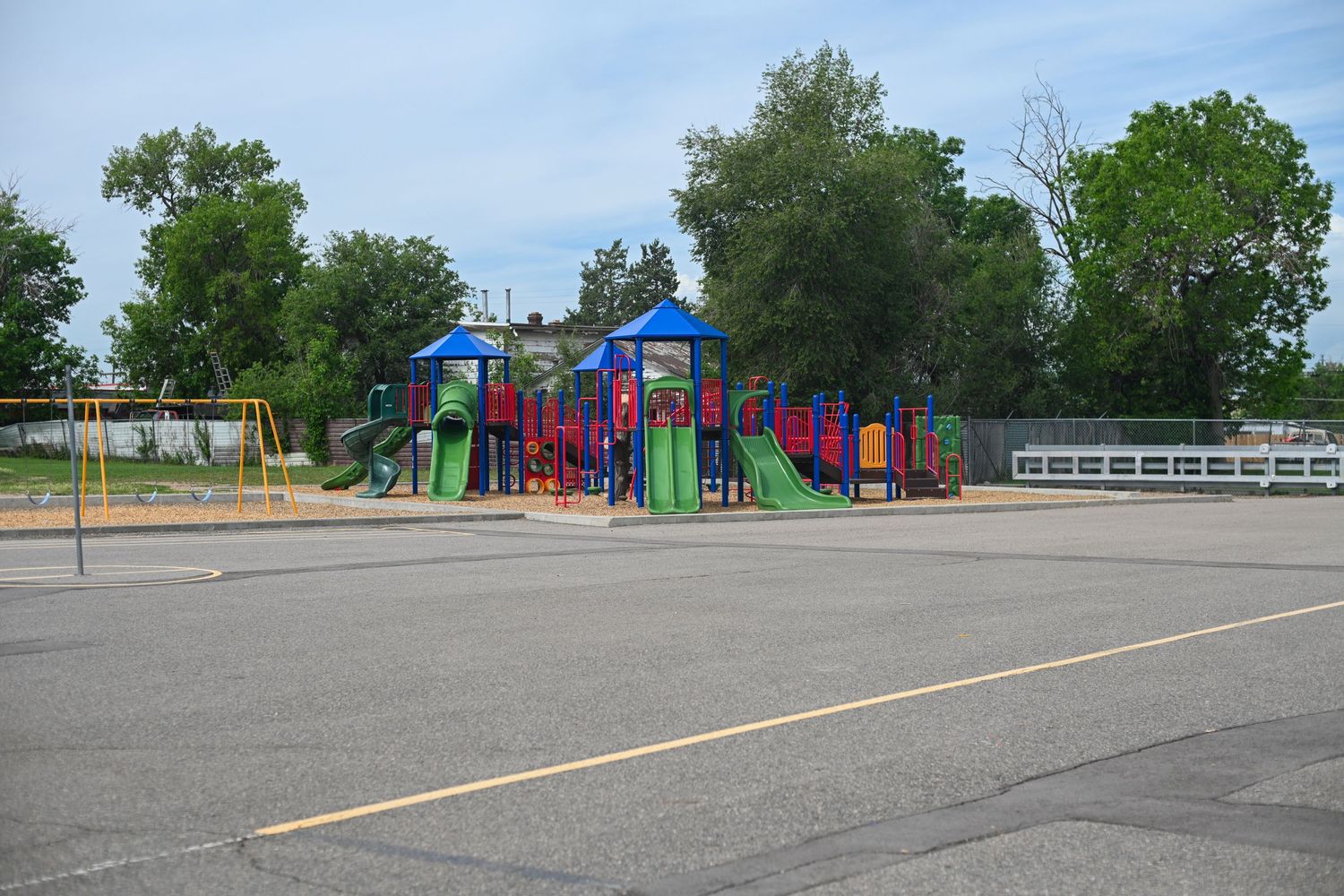 Colorful playground equipment with slides and climbing structures in an empty school parking lot surrounded by trees.