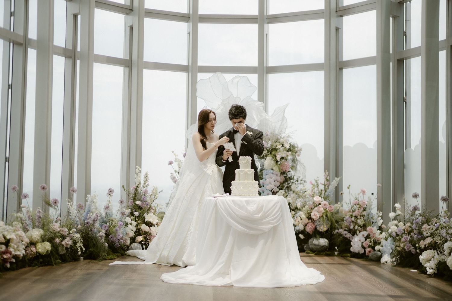 Bride and groom cut their wedding cake surrounded by floral arrangements and large windows.
