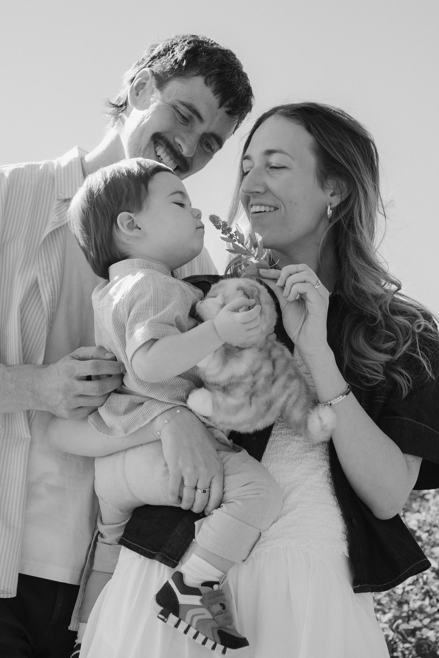 B&W family laughing as toddler sniffs a flower in family lifestyle photo session | Candid family photography, North Vancouver
