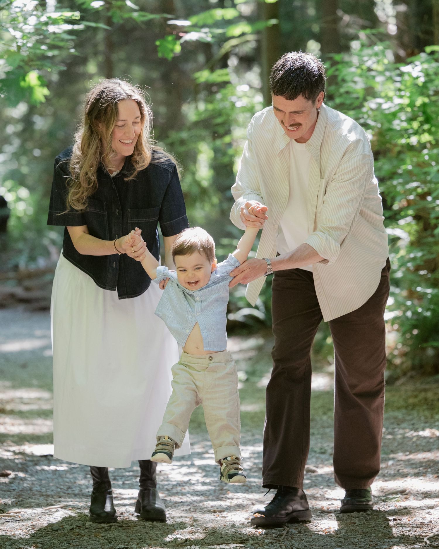 Parents swinging their toddler between them on a forest path at Lighthouse Park, natural family photography in West Vancouver Tashtakespics