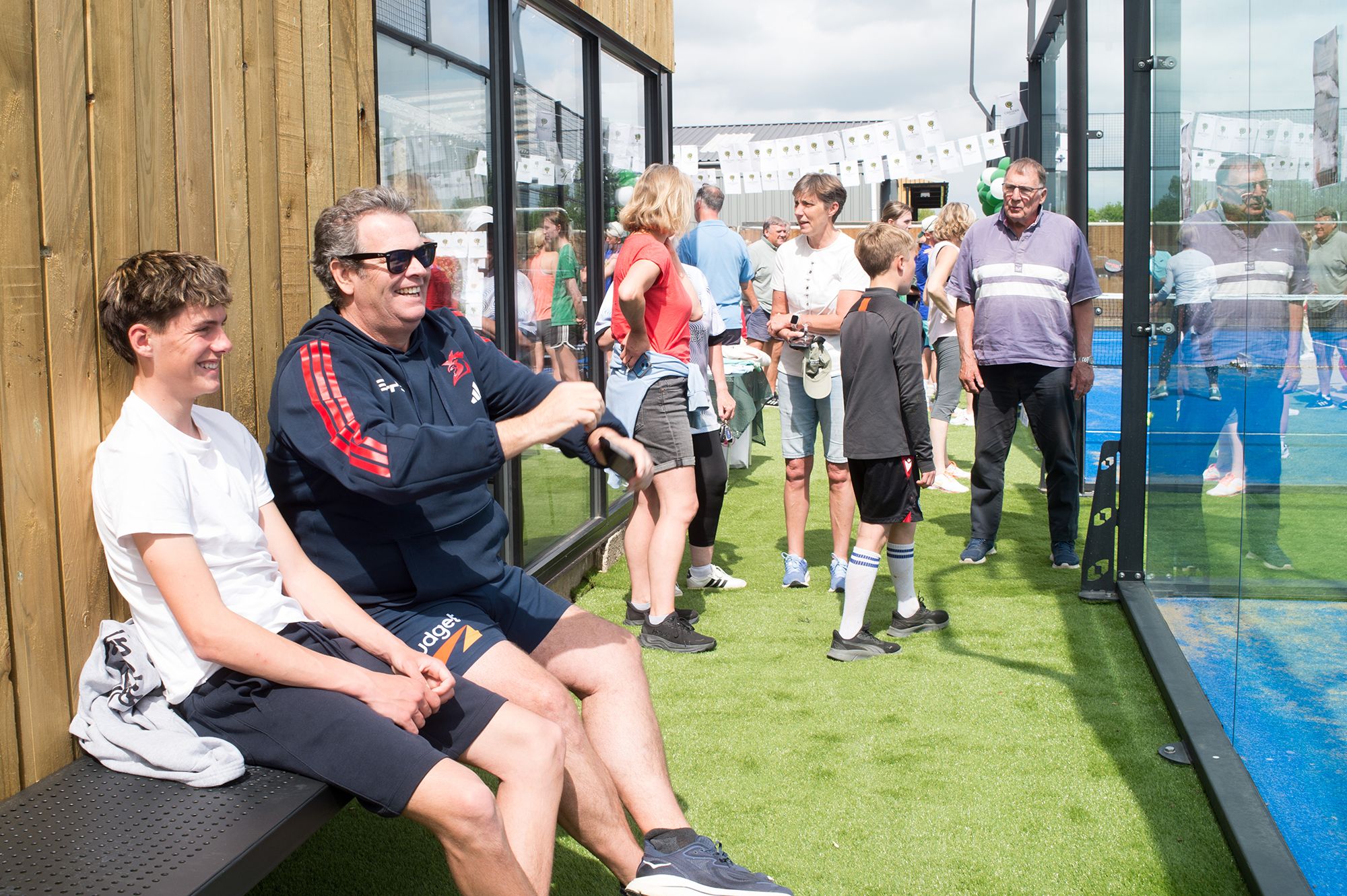 A man sits on a bench surrounded by a group of people at the Chiltern Padel launch party in Buckinghamshire.
