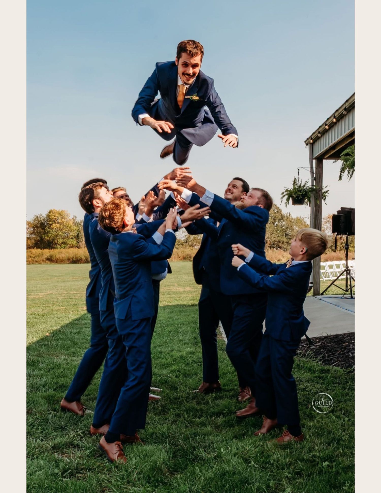 Groomsmen in navy suits tossing groom into the air outdoors at a wedding celebration on a sunny day.