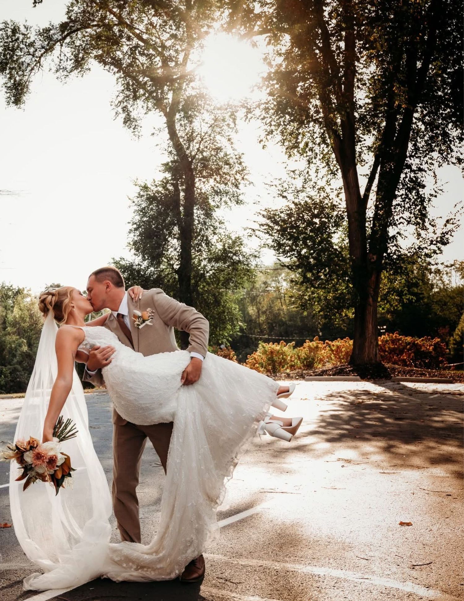 Groom dips bride for a romantic kiss outdoors, sunlight filtering through trees, bride holding bouquet in white gown.