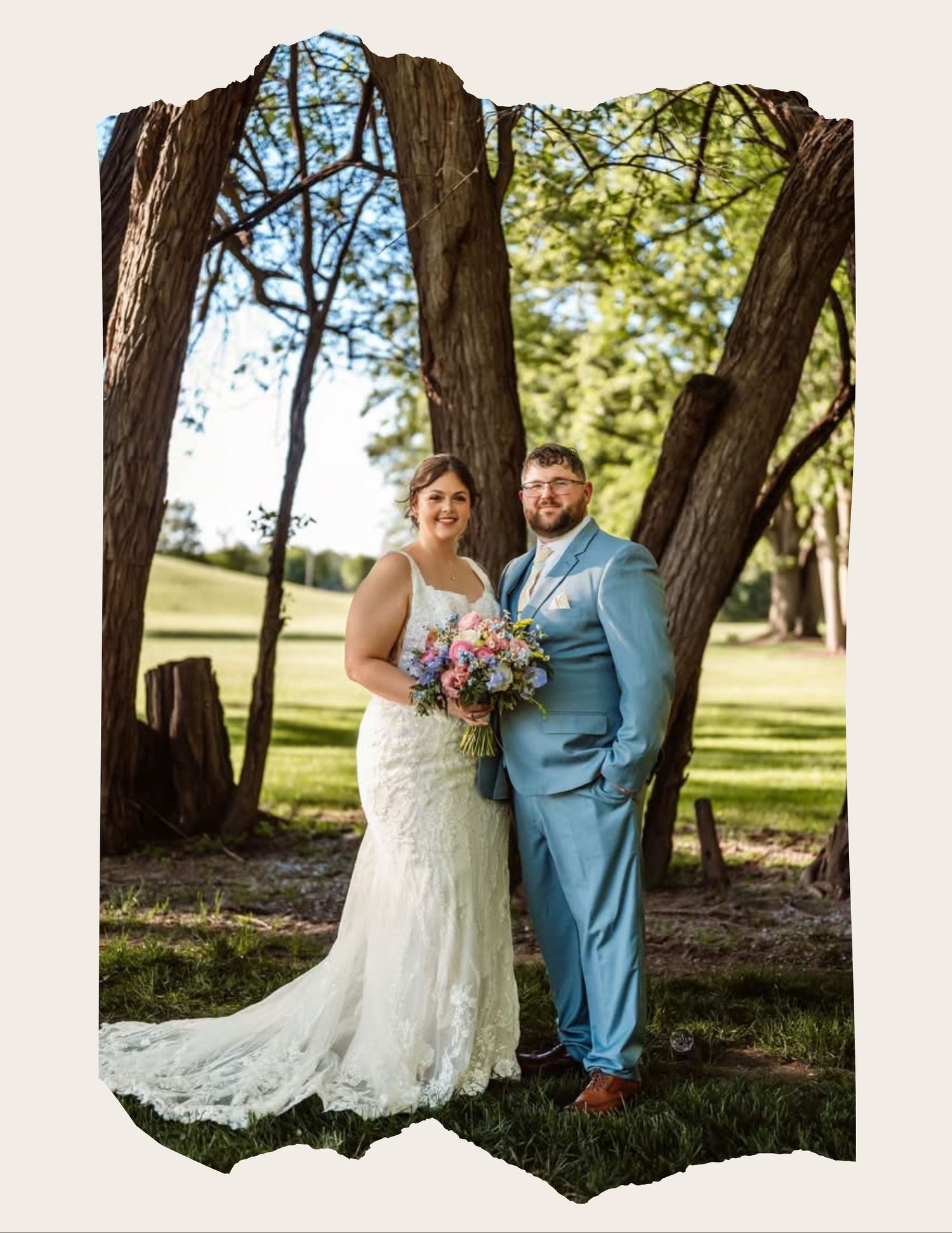Bride in lace gown and groom in light blue suit pose together outdoors under trees on their wedding day.