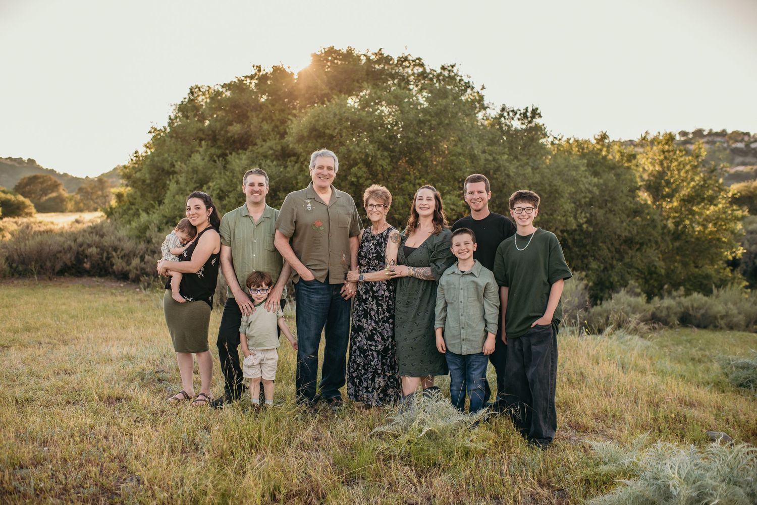 Multi-generational family portrait outdoors in golden sunset light, standing together in a green meadow with trees behind them.
