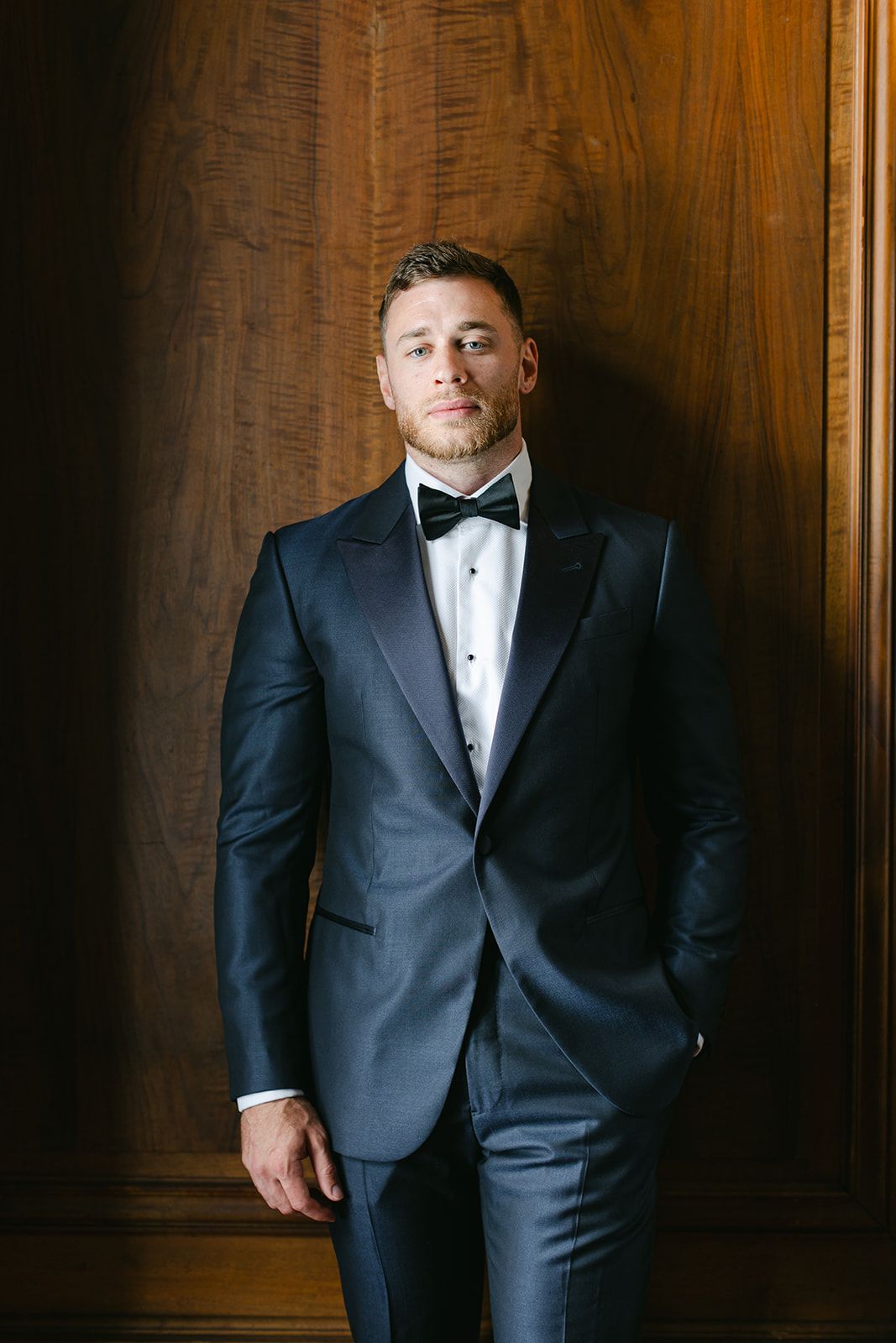 Armour House Wedding - Groom in navy tuxedo with black bow tie poses confidently against a rich wooden wall.
