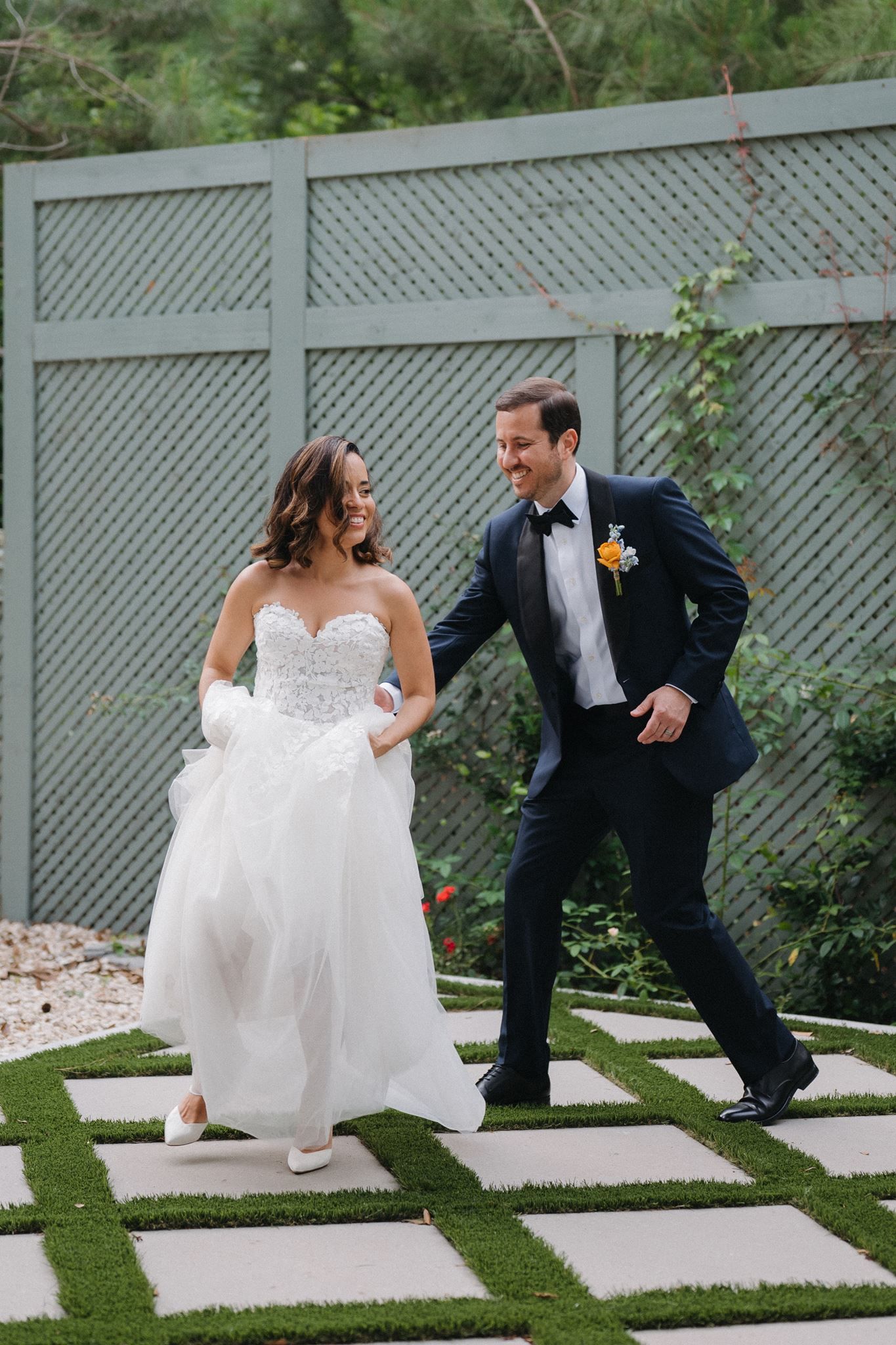 Joyful bride and groom in wedding attire walking together outdoors on a patterned stone path surrounded by greenery.