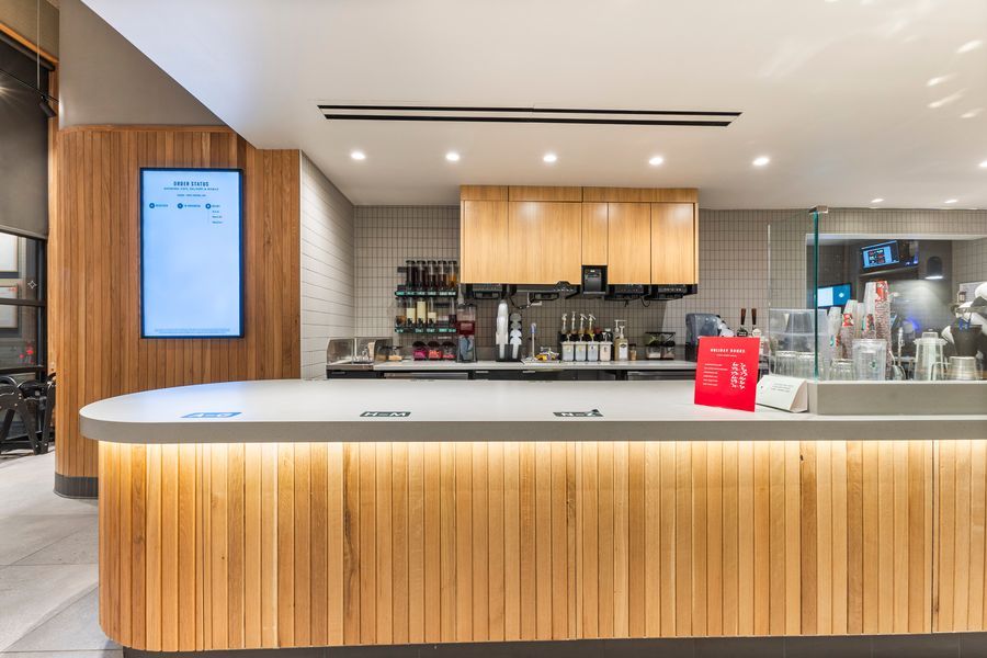 Modern airport lounge bar with warm wood paneling, curved counter, wine bottles, and recessed lighting.