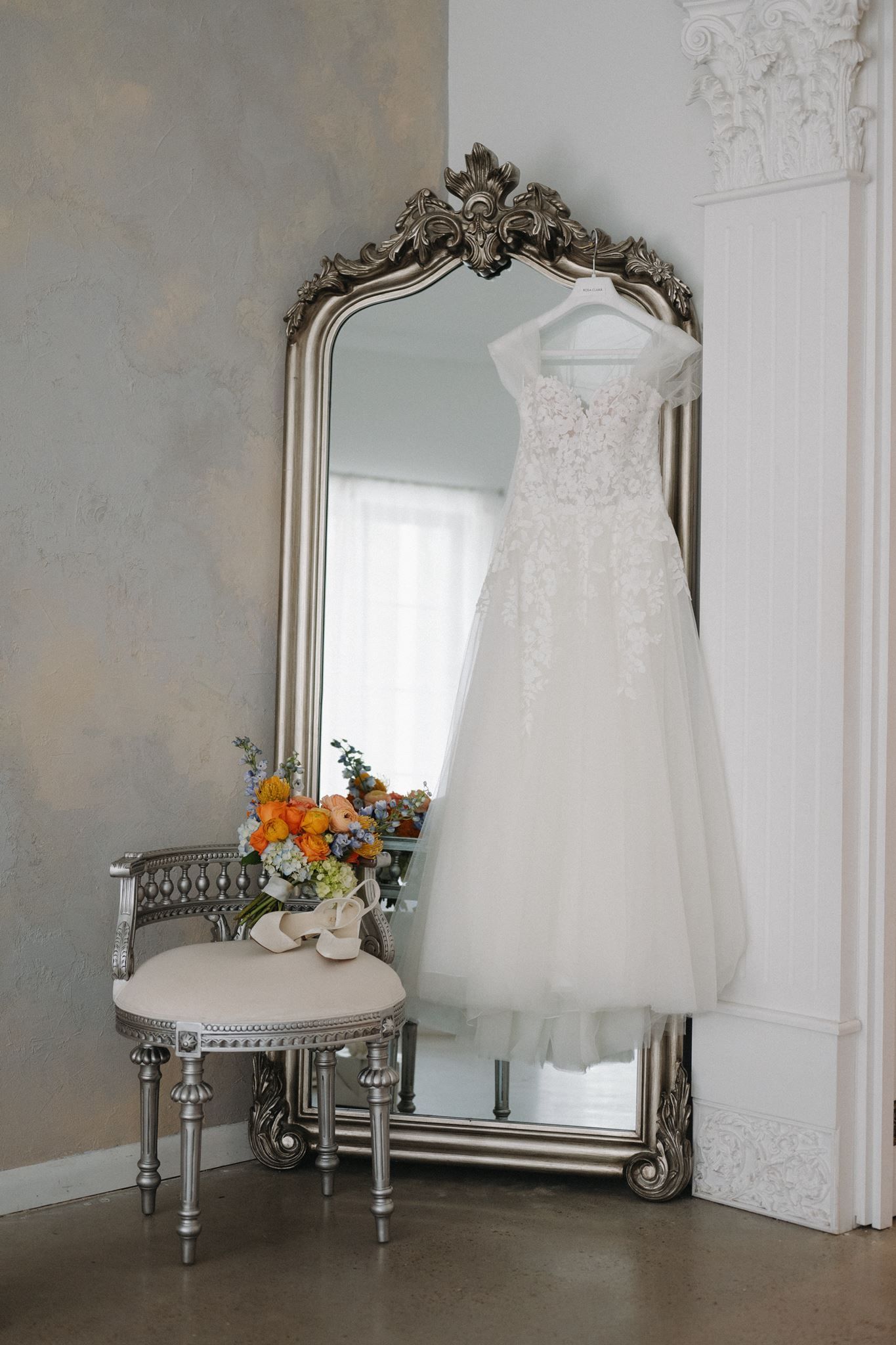 Elegant white lace wedding dress hanging on ornate silver mirror beside floral bouquet on vintage chair.