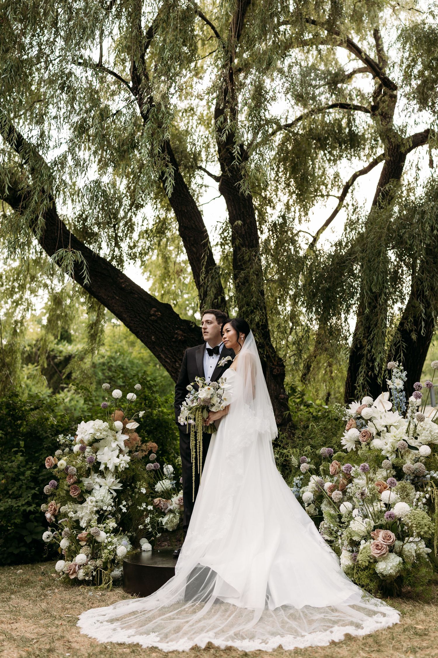 Bride and groom pose under a willow tree surrounded by elegant floral arrangements on their wedding day.
