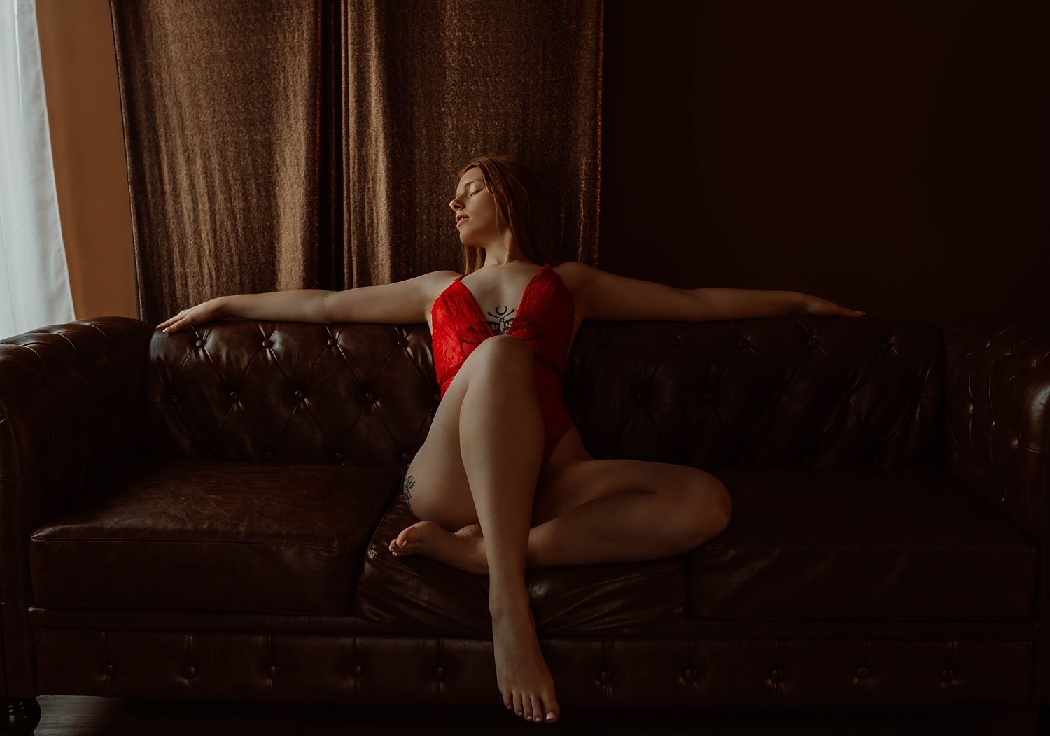 redhead in red bodysuit lounges over leather couch by KJ Boudoir Photography