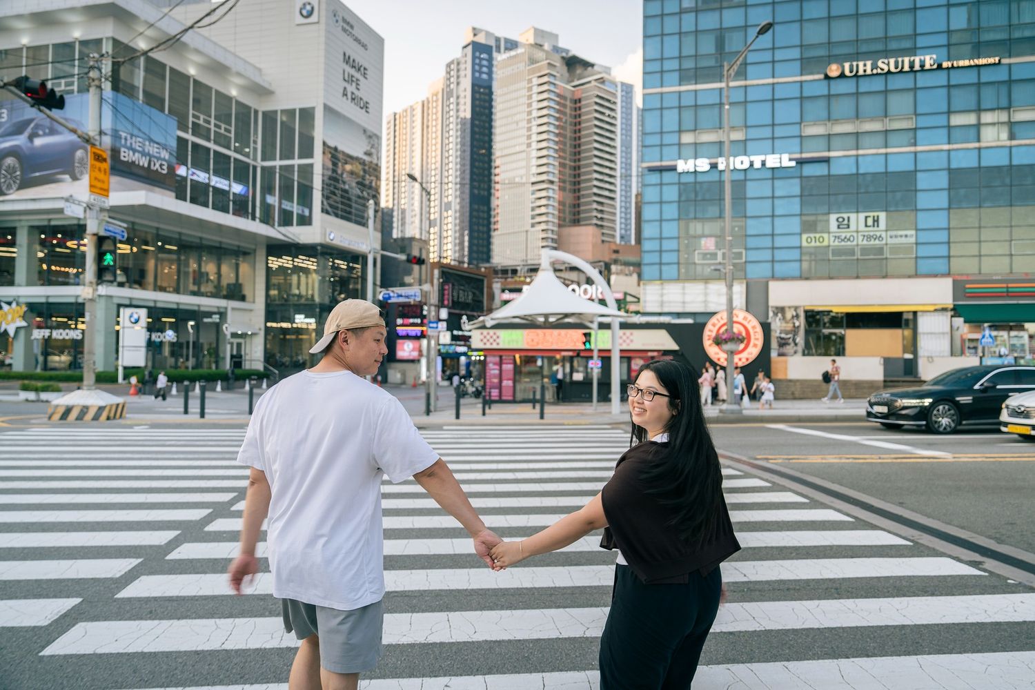 Couple holding hands crossing a busy urban intersection with city buildings and a hotel in the background.