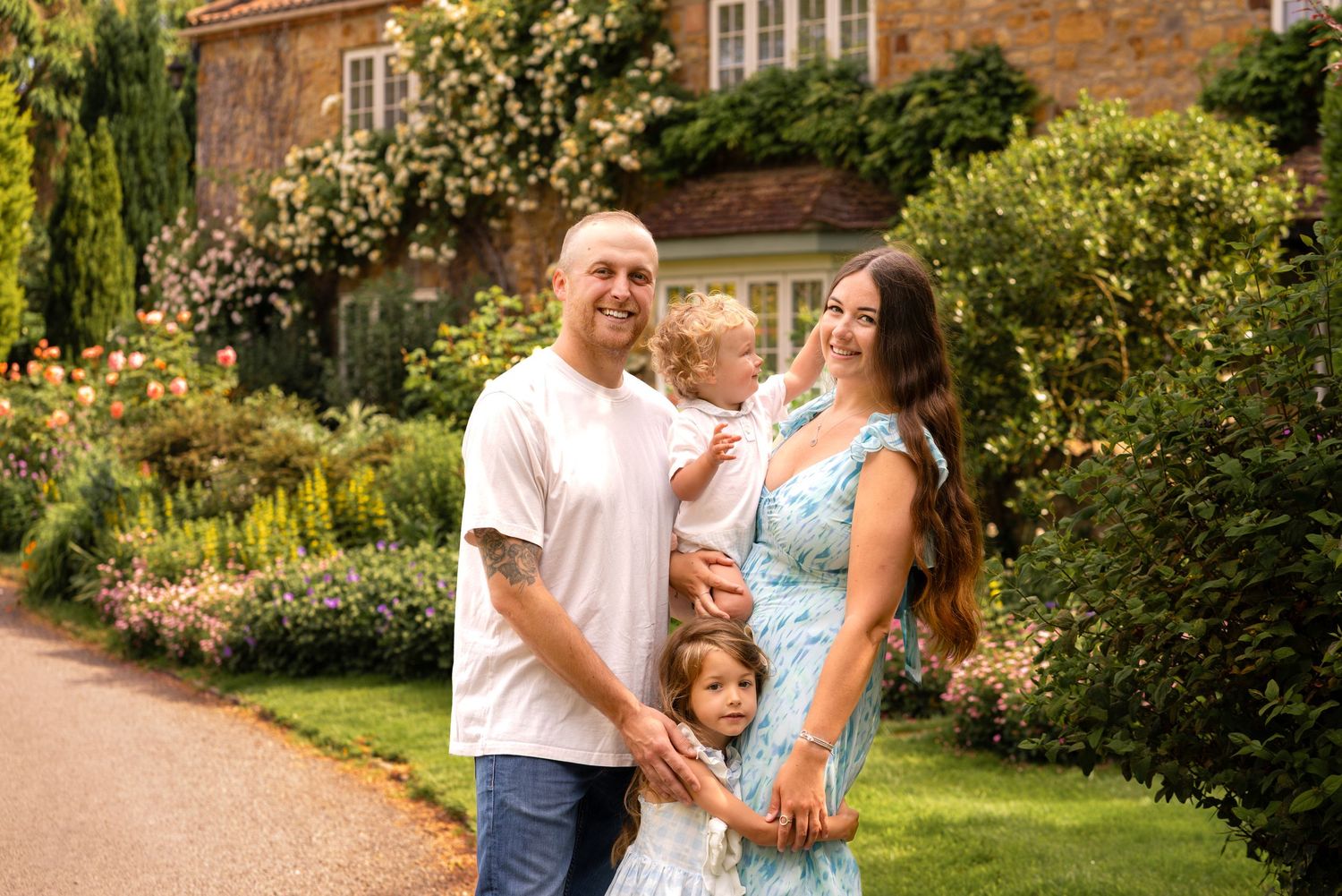 Happy family of four posing in a lush garden with blooming flowers and a brick house backdrop on a sunny day.