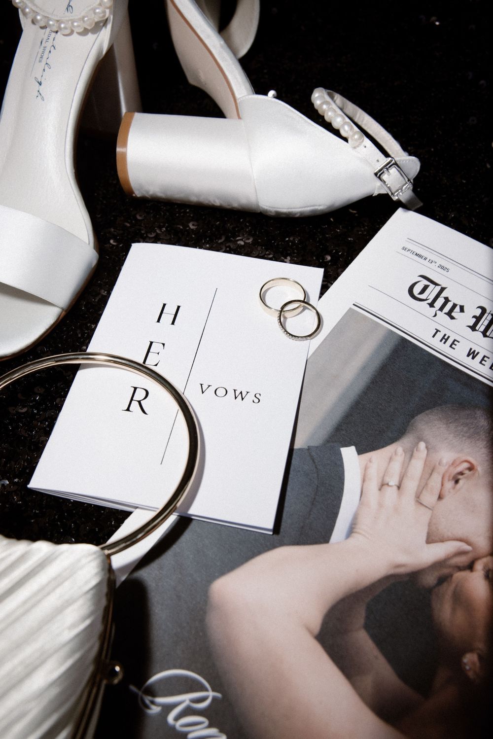 Elegant wedding flat lay with white heels, vow books, gold rings, pearl clutch, and newspaper on dark background.