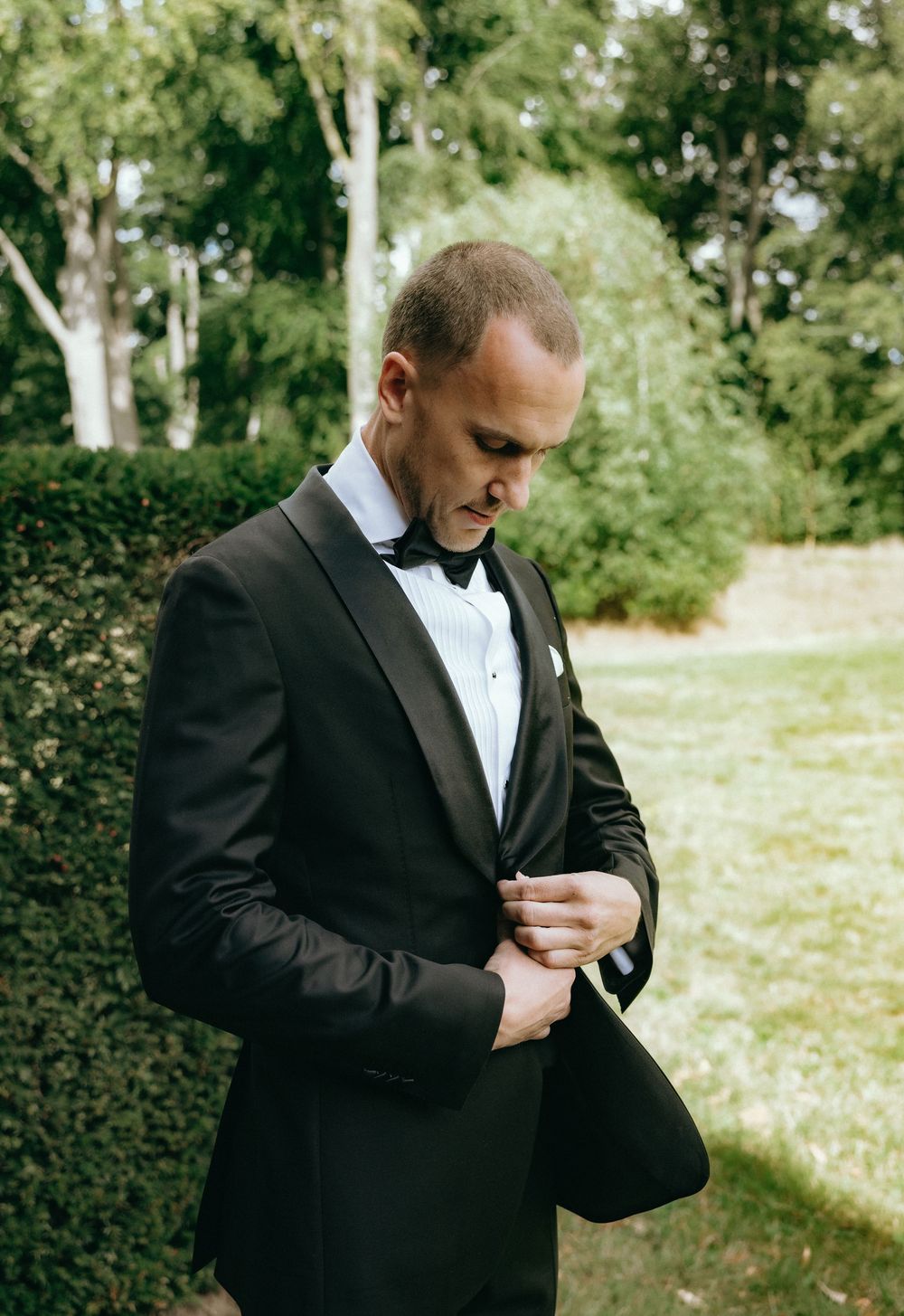 Groom in black tuxedo adjusting jacket outdoors with lush green hedges and trees in background.