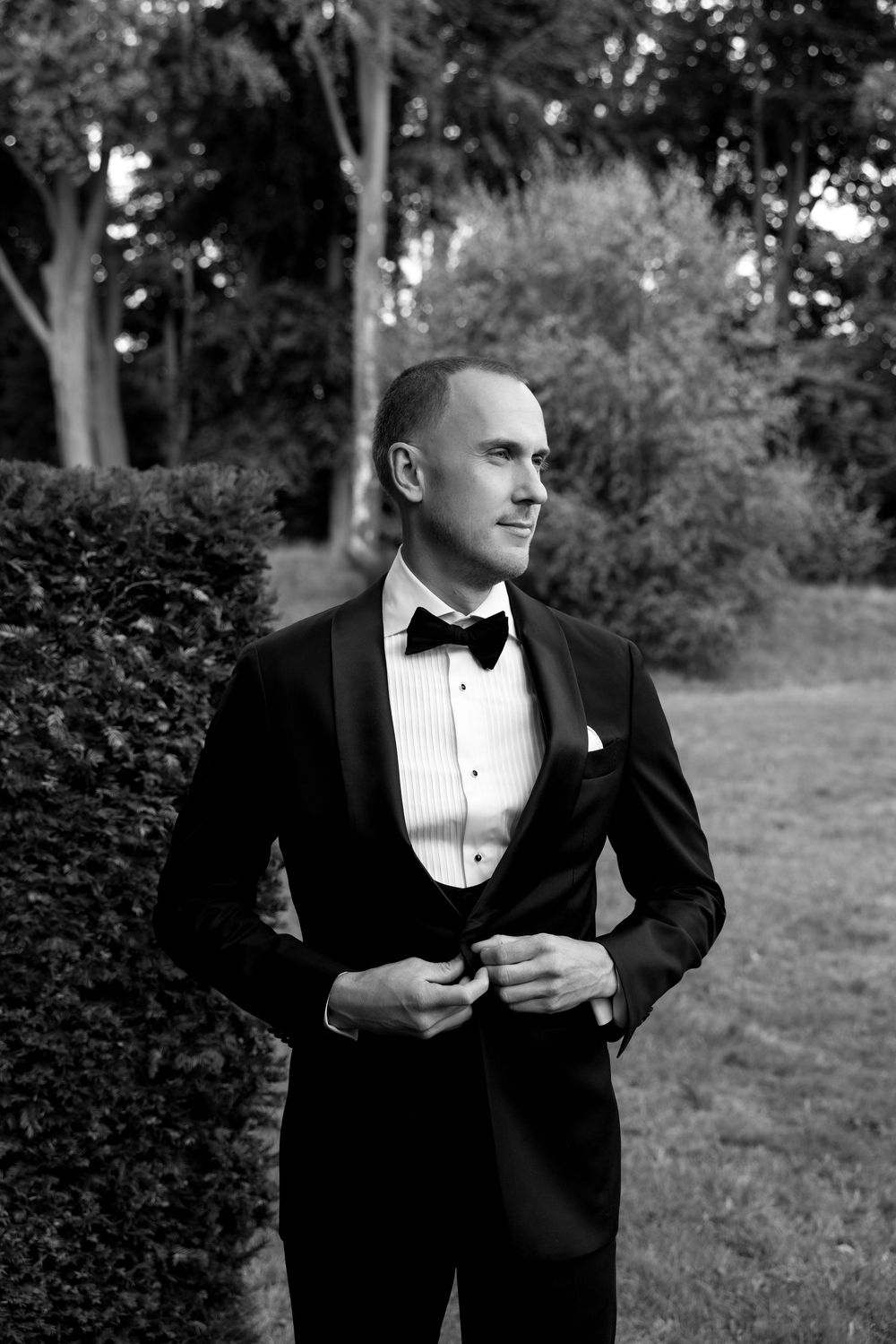 Groom in black tuxedo with bow tie buttoning jacket outdoors in garden, elegant black and white wedding portrait.