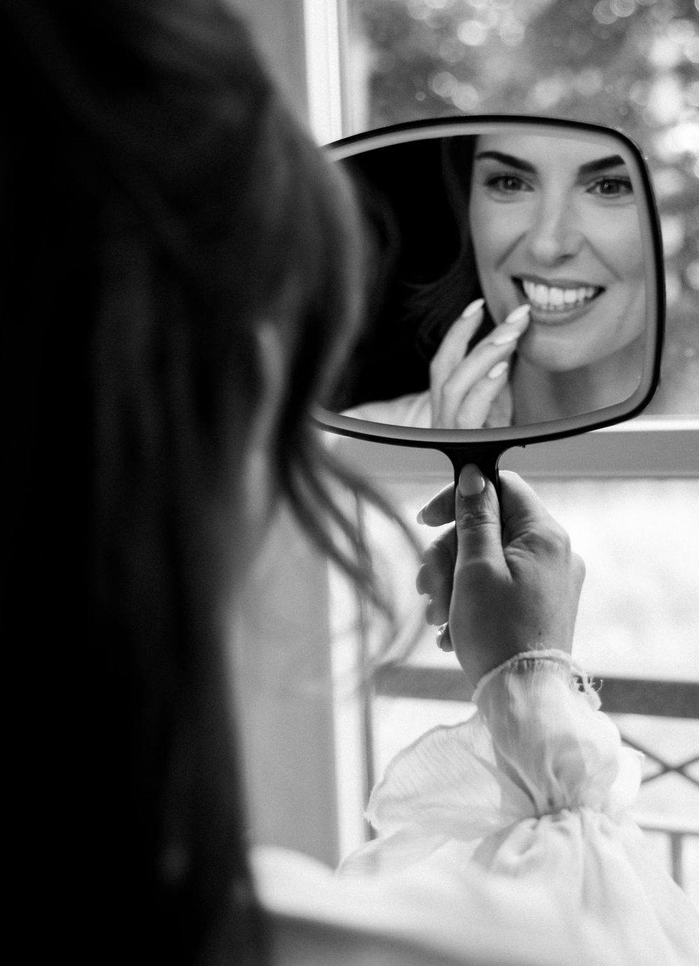 Black and white photo of a smiling woman applying lipstick while looking into a handheld mirror.