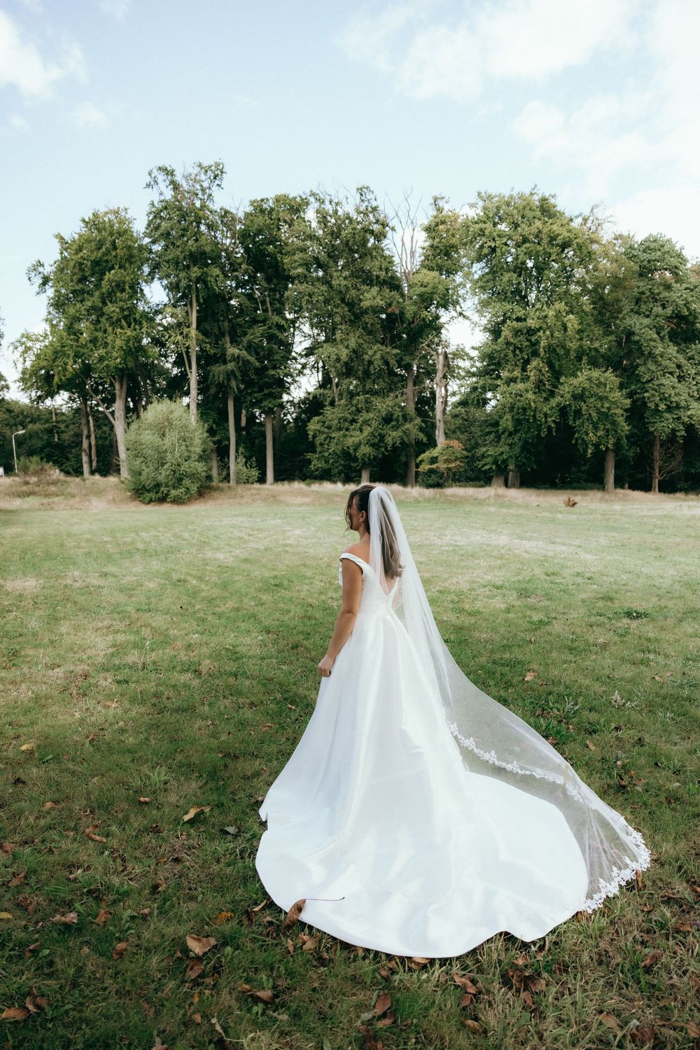 Bride in elegant white gown with long train and veil stands in lush green park on wedding day.