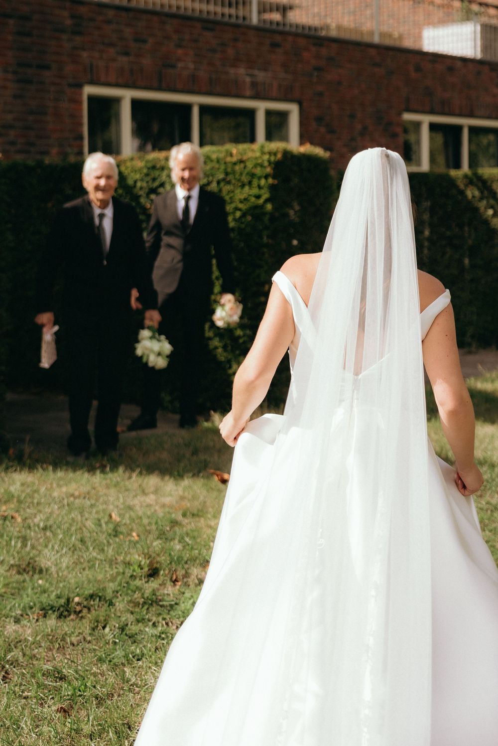 Bride in white gown and veil walks toward two men in suits outside a brick building on a sunny wedding day.