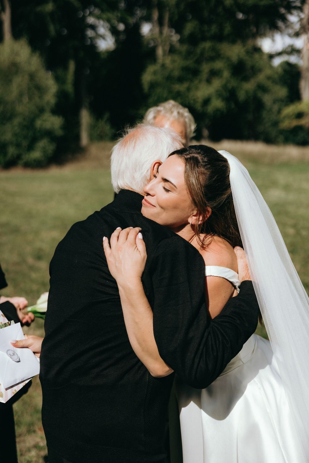 Bride in white gown and veil sharing an emotional hug with an older guest at an outdoor wedding ceremony.