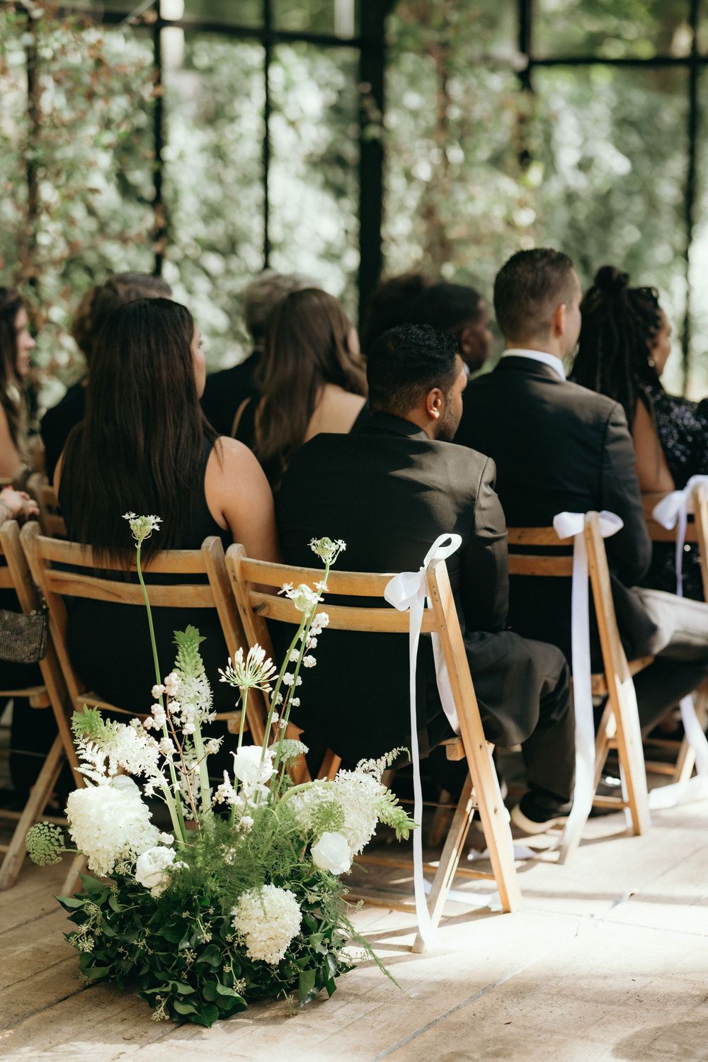Wedding guests seated in wooden folding chairs during an outdoor ceremony with white floral arrangements.