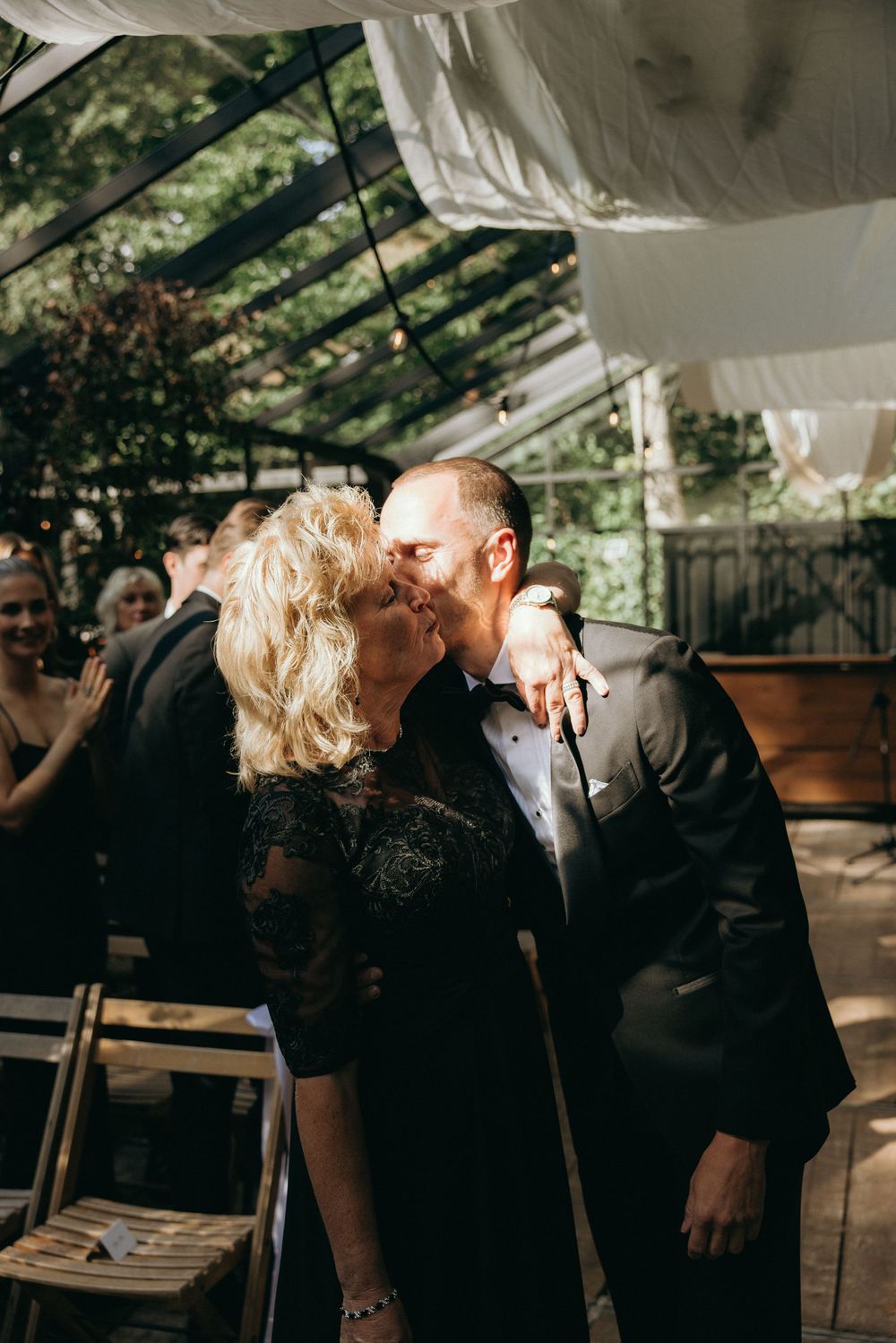 Groom sharing a kiss with older woman in black lace dress during wedding ceremony under glass canopy.