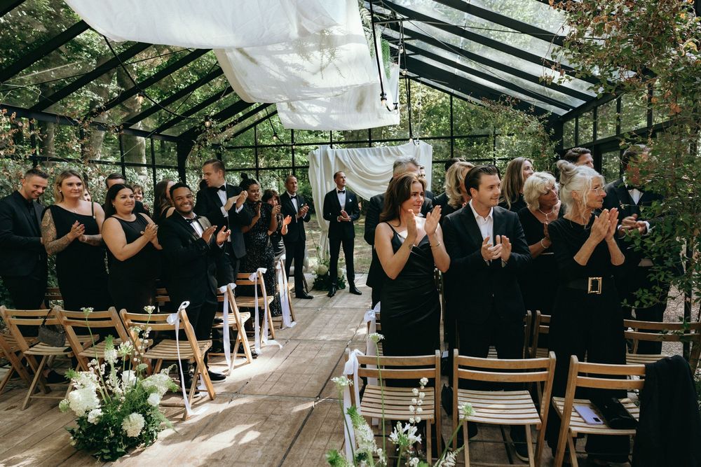 Guests in black attire applaud during an elegant greenhouse wedding ceremony with wooden chairs and floral decor.