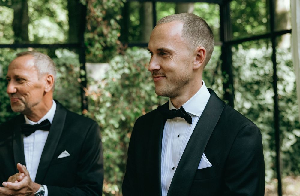 Two men in black tuxedos with bow ties stand outdoors, smiling at a formal wedding ceremony.