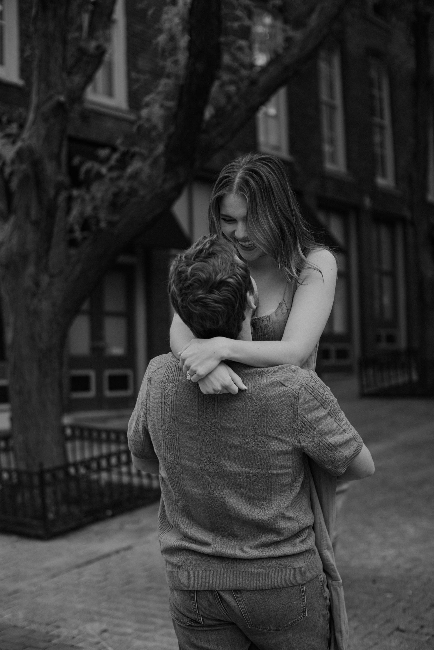 A woman smiles down at her fiance as he lifts her into the air and spins her around in front of a brick building.