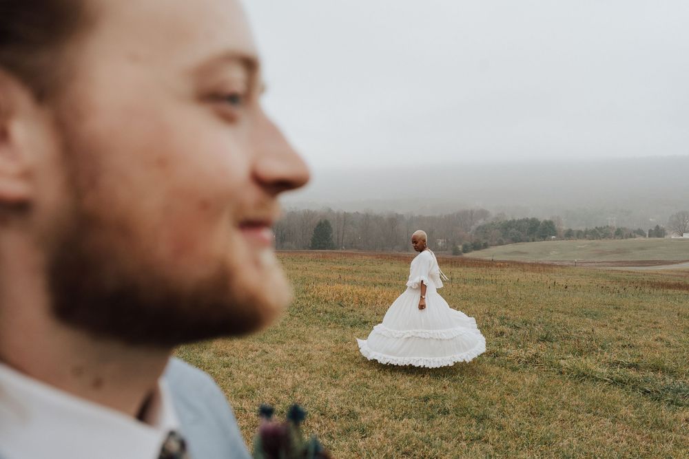 A groom smiles in the foreground as his bride in a white wedding gown stands in a misty green field.