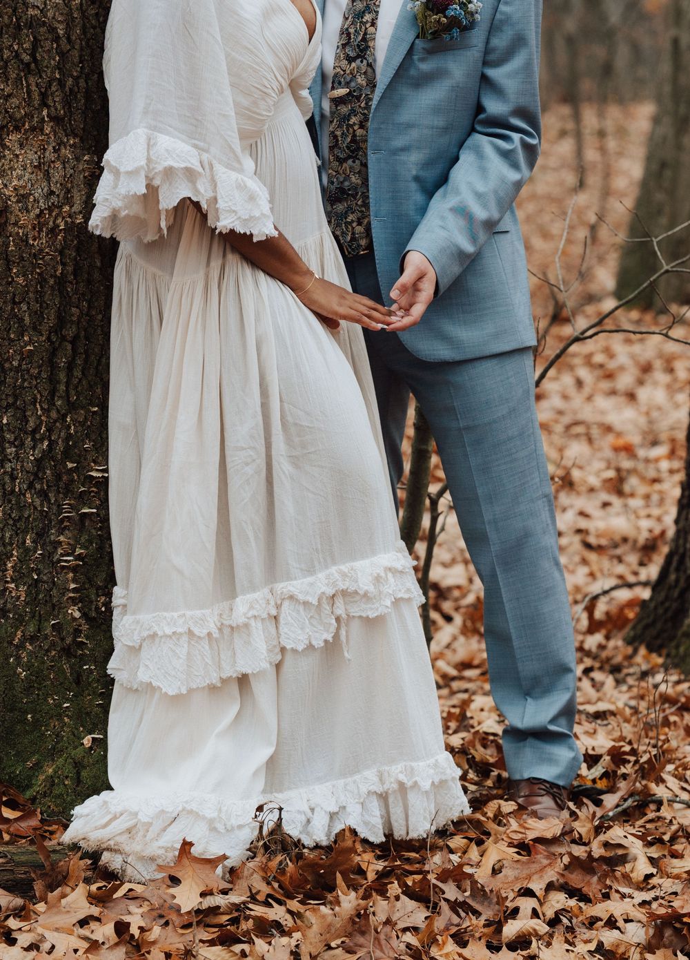 Bride in ruffled white boho gown holds hands with groom in light blue suit among autumn leaves in forest.