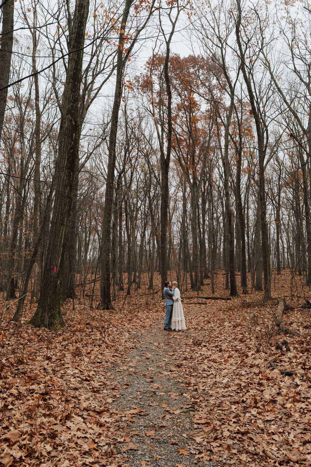 A couple embraces on a leaf-covered forest path surrounded by bare autumn trees on an overcast day.