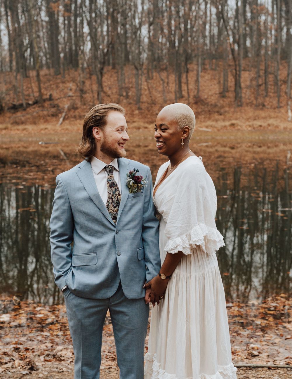 A couple in wedding attire holds hands by a pond in autumn, smiling at each other.