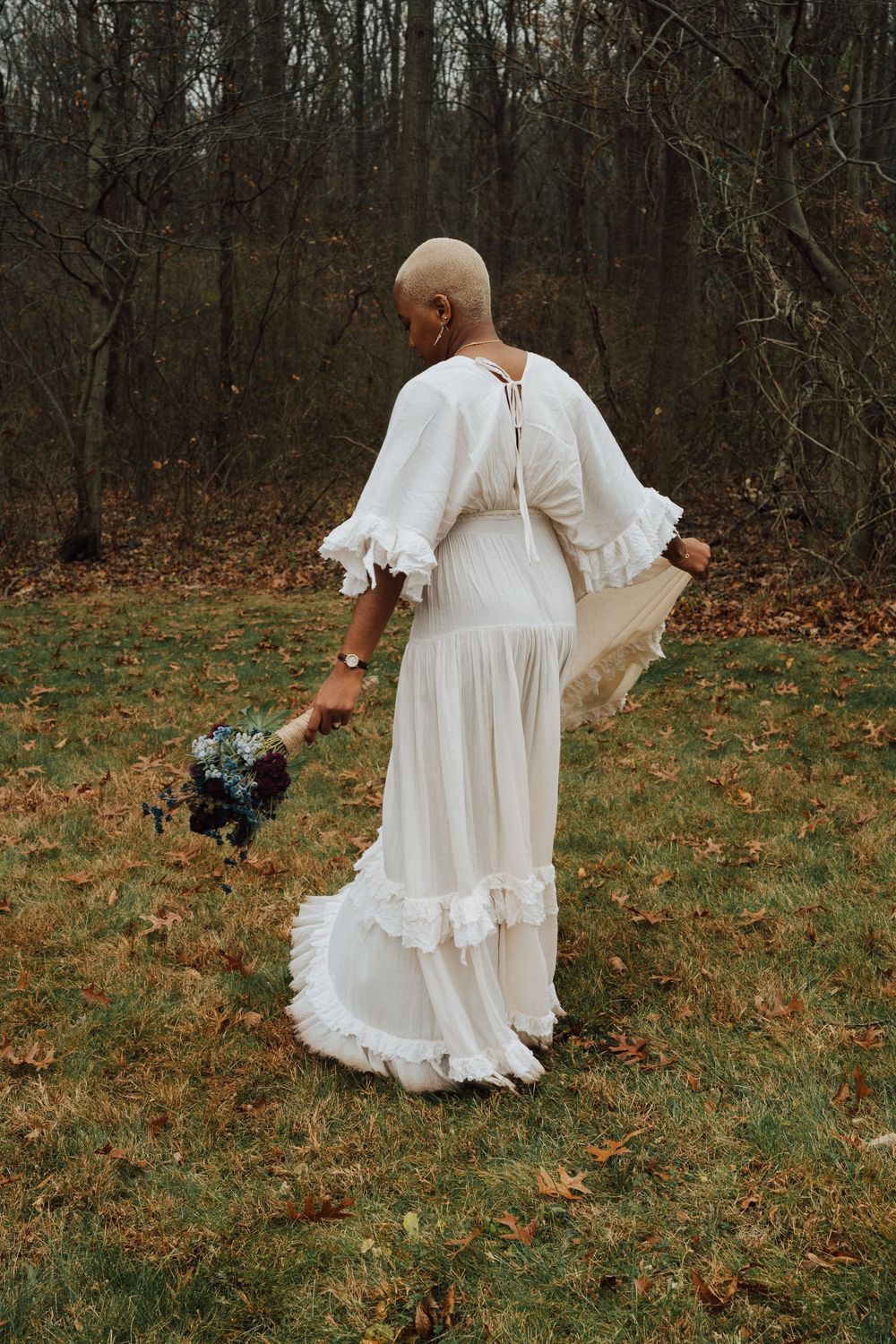 Bride in white ruffled boho gown holding wildflower bouquet in autumn forest clearing, viewed from behind.