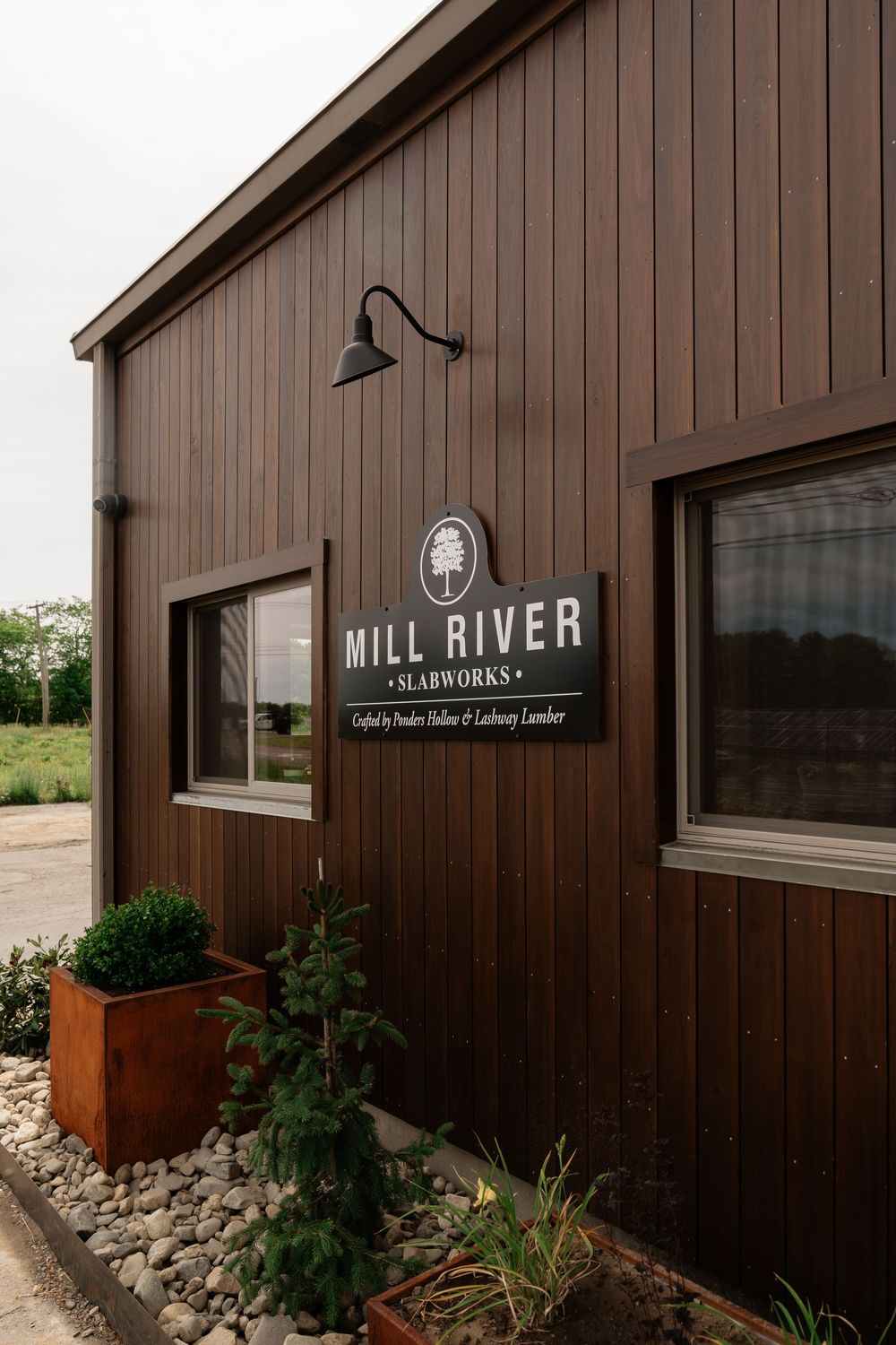 Mill River Slabworks exterior building sign on dark brown wood siding with gooseneck light and landscaping.
