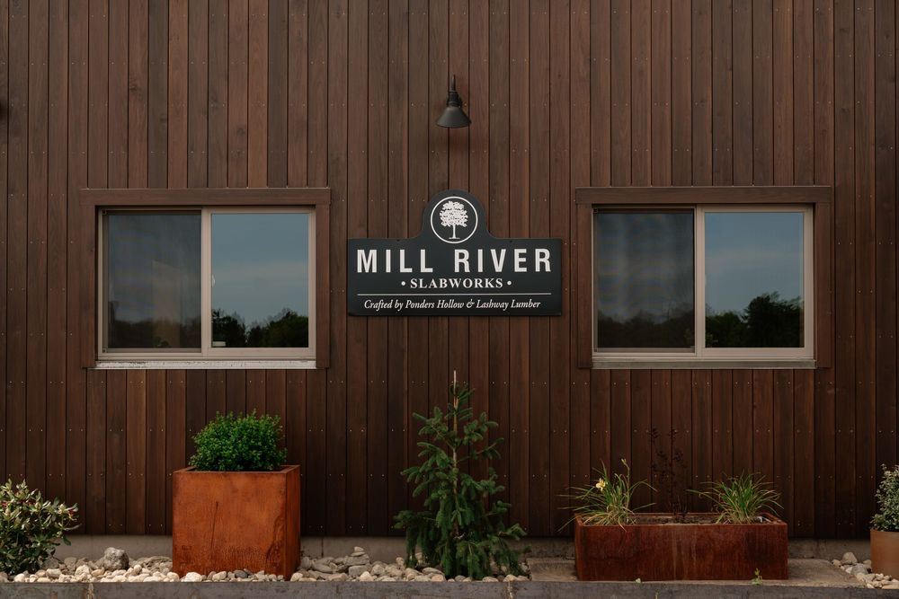 Exterior of Mill River Slabworks building with wooden siding, signage, and decorative planters with greenery.