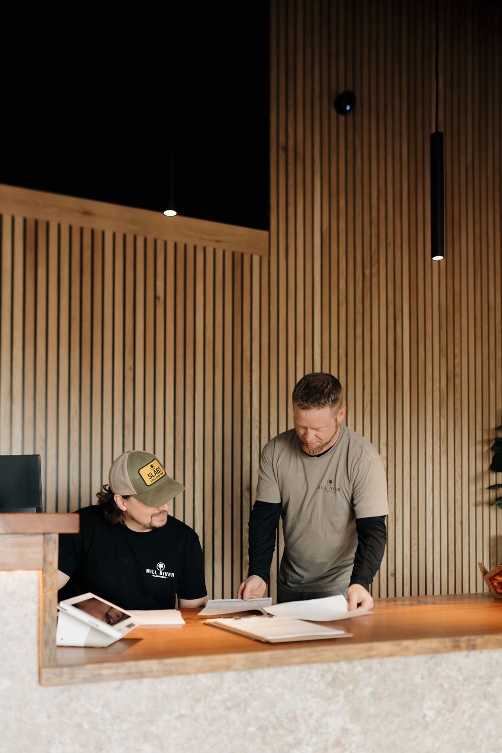 Two employees reviewing documents at a wooden reception desk with slatted wood wall backdrop.