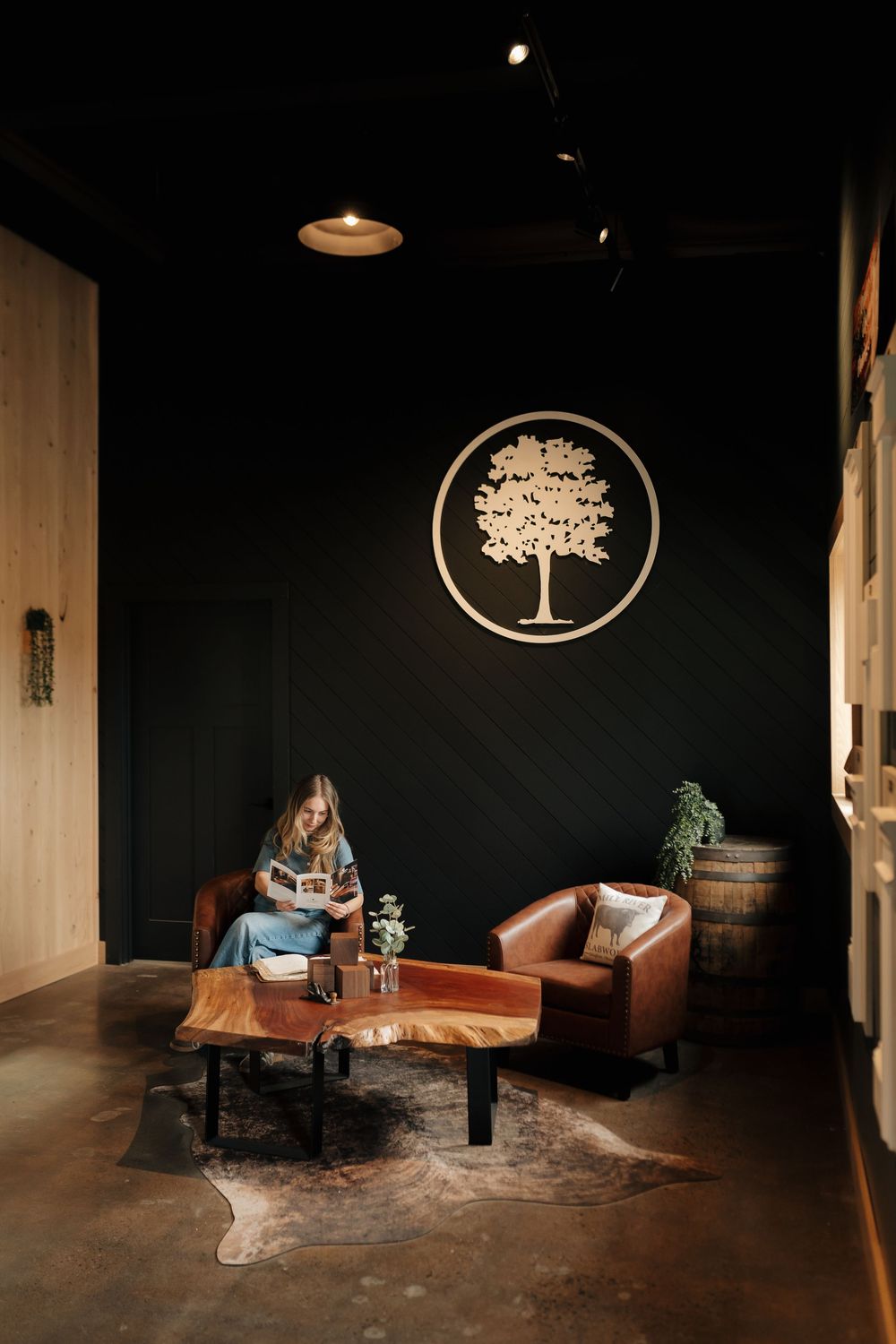 Woman reading in a rustic lounge with leather chairs, live-edge table, and decorative tree logo on dark wall.